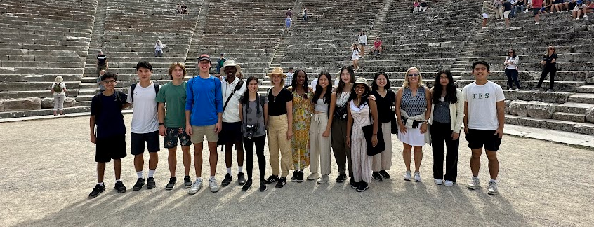 A group shot of about a dozen students surrounded by the ruins of the Epidaurus, the birthplace of the healing god, Asclepios.