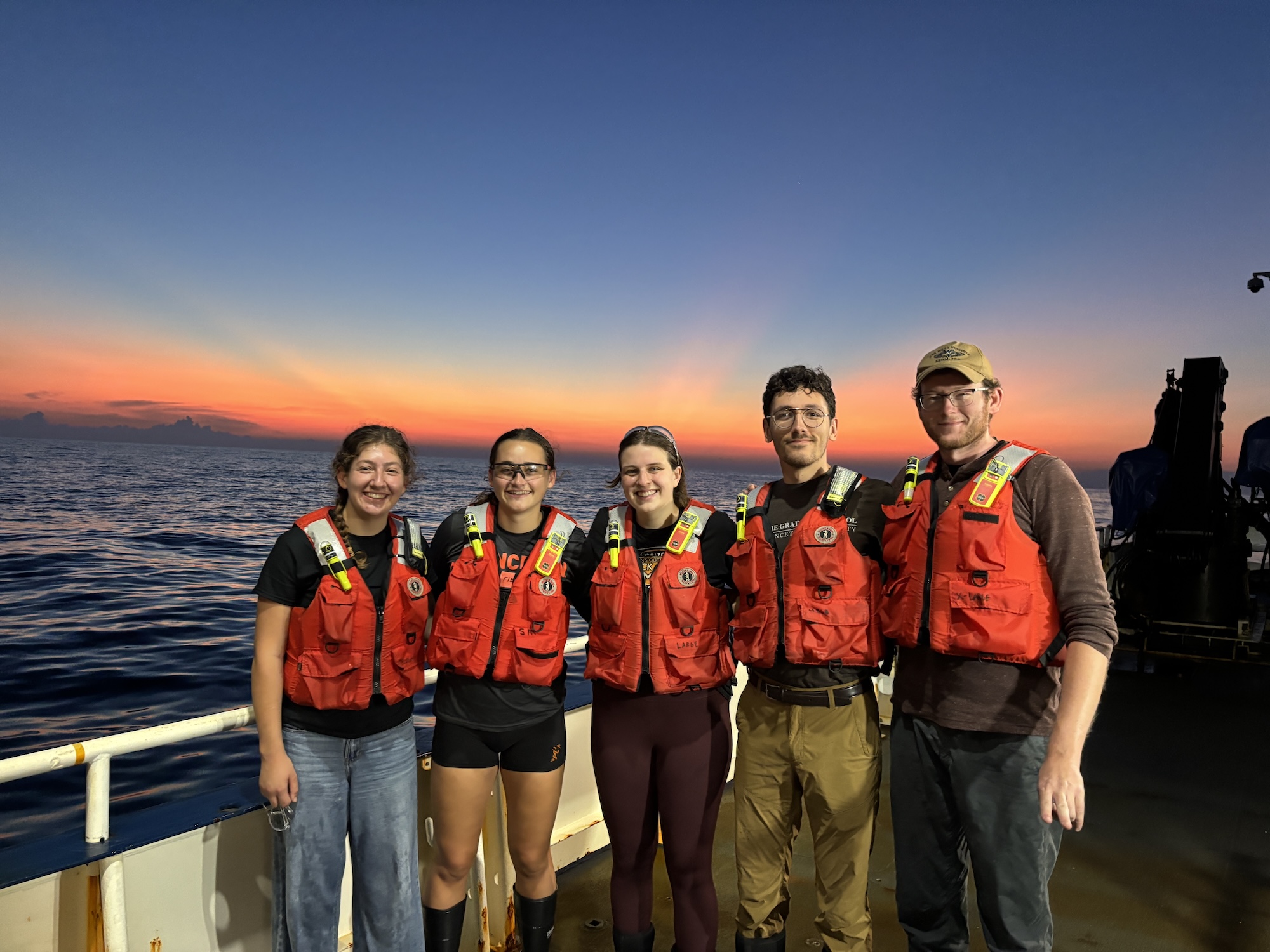 5 students wearing red life jackets on boat with sunset in background