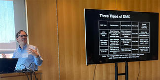 A man gives a presentation at a lectern with a flat screen filled with data behind him