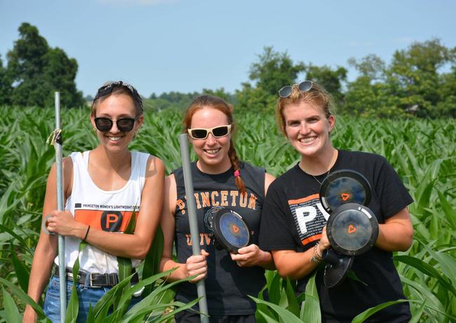 Three students in t-shirts pose and smile for camera while holding light farming equipment. They are standing in a crop field.