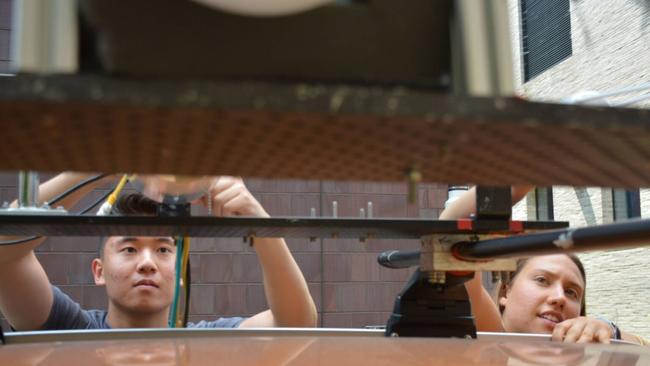 Two students reach up to a rack of electronics installed on the external roof of a car. Both students are concentrated on their work.