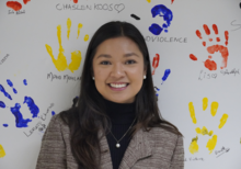 A female student stands in front of a large canvas with children's hand prints in paint.