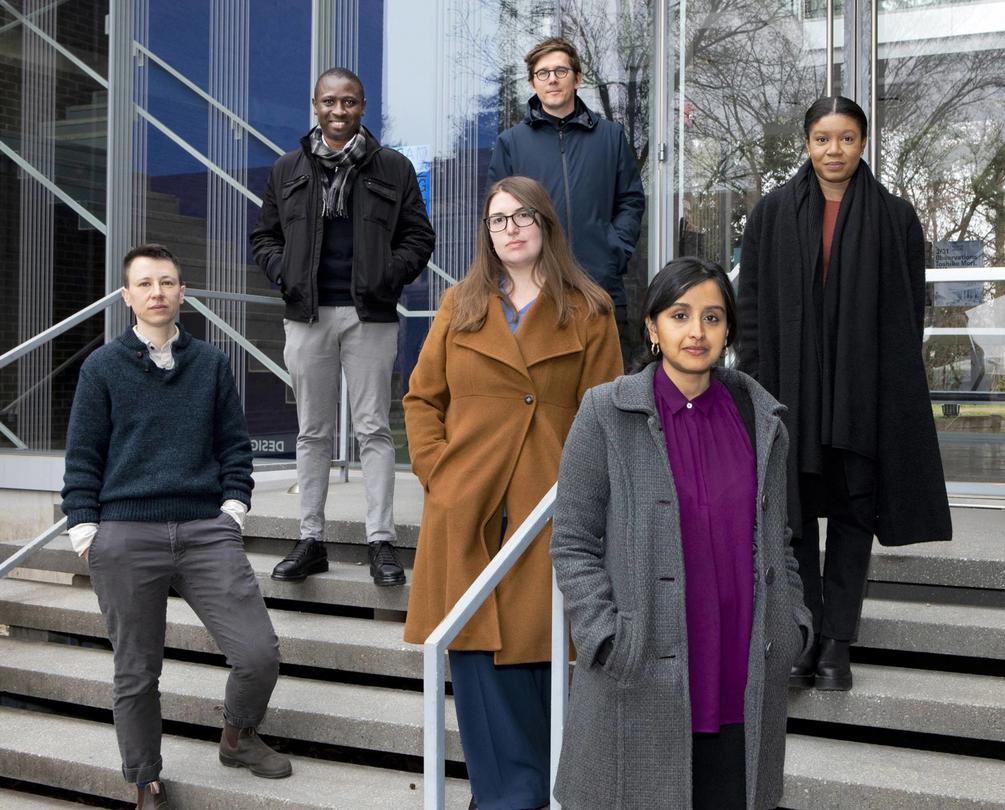 Six smiling students in winter coats, standing on the steps of a campus building.