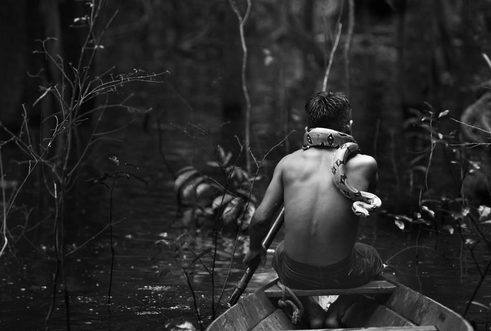A black and white photograph of a young person navigating a small boat down the Solimões River in the Brazilian Amazon. The person has a snake wrapped around their neck.