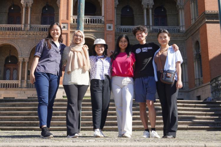 Six students stand with their arms around each other in front of a building.
