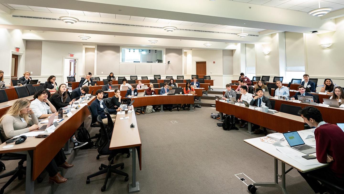 Students sit in a large seminar ropm with long tables