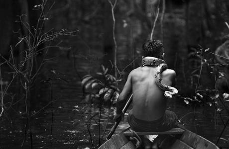 A black and white photograph of a young person navigating a small boat down the Solimões River in the Brazilian Amazon. The person has a snake wrapped around their neck.