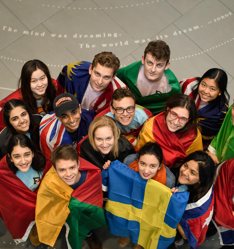 A group of students look up a from the Louis A. Simpson Atrium while draped in the flags of their home countries. 